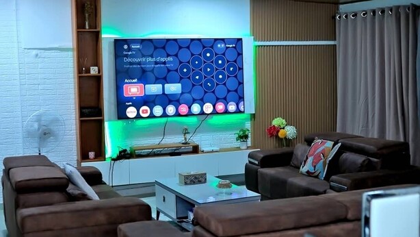 Una sala de estar moderna con un televisor de pantalla plana grande montado en una pared blanca, un sofá marrón, una mesa de centro y una mesita auxiliar con adornos.