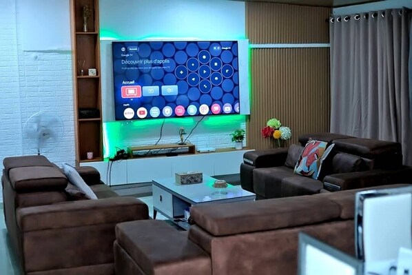 Una sala de estar moderna con un televisor de pantalla plana grande montado en una pared blanca, un sofá marrón, una mesa de centro y una mesita auxiliar con adornos.