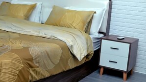 Una cama bien hecha con un edredón estampado y almohadas, una mesita de noche con cajones y una pared de ladrillo blanco al fondo.
