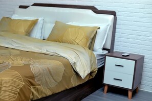 Una cama bien hecha con un edredón estampado y almohadas, una mesita de noche con cajones y una pared de ladrillo blanco al fondo.