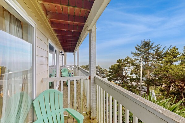 A wooden balcony with white railings, a green chair, and a view of trees and the ocean. - Easy Access Dog Friendly Studio with Ocean Views Short Walk to Beach (Lincoln City)