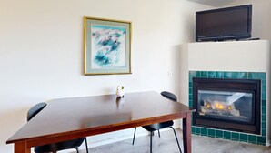 Un coin repas avec une table et des chaises en bois, une cheminée agrémentée de carreaux de céramique, un tableau abstrait encadré accroché au mur, et un téléviseur à écran plat fixé au-dessus de la cheminée.