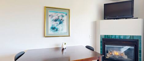 Un comedor con mesa y sillas de madera, una chimenea con revestimiento de azulejos, un cuadro abstracto enmarcado en la pared y un televisor de pantalla plana montado sobre la chimenea.