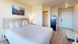 Une chambre d’hôtel avec un grand lit, une table de chevet avec une lampe, un réfrigérateur, une porte et une petite table avec une chaise.