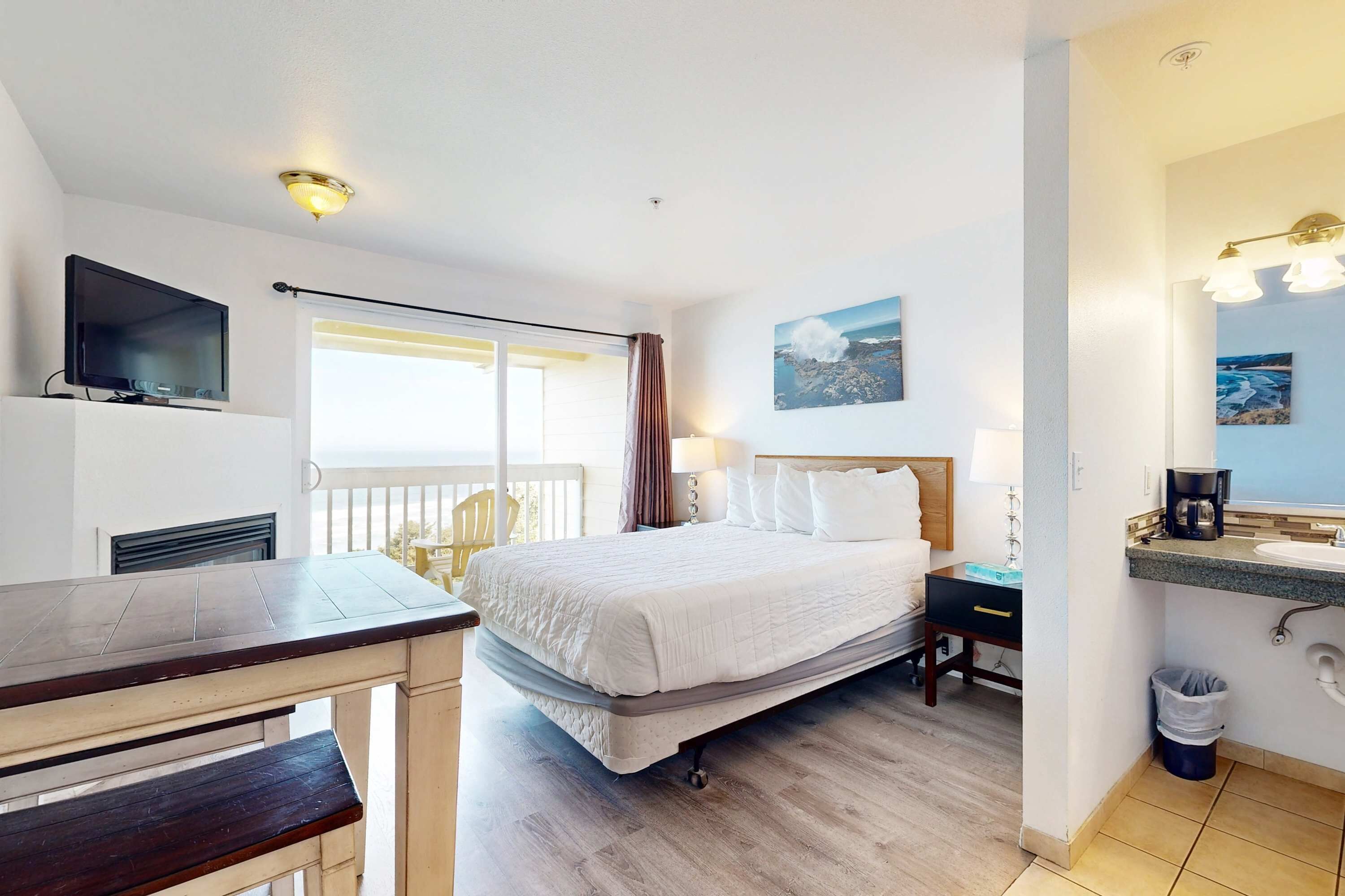 A hotel room with a bed, a desk, a TV, a balcony, and a bathroom with a sink and mirror.