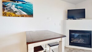 A room with a white wall, a flat-screen TV mounted above a fireplace, and a painting of a coastal scene. - Upper Floor Oceanview Studio Perfect for Couples Free WiFi Dogs Allowed (Lincoln City)