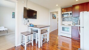 Una cocina moderna con armarios de madera, microondas integrado, estufa y refrigerador. Hay un pequeño comedor con una mesa y dos taburetes.