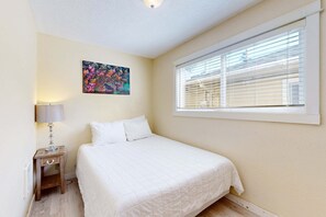 Un dormitorio con cama, una mesita de noche con lámpara, una ventana con persianas y un cuadro de colores en la pared.
