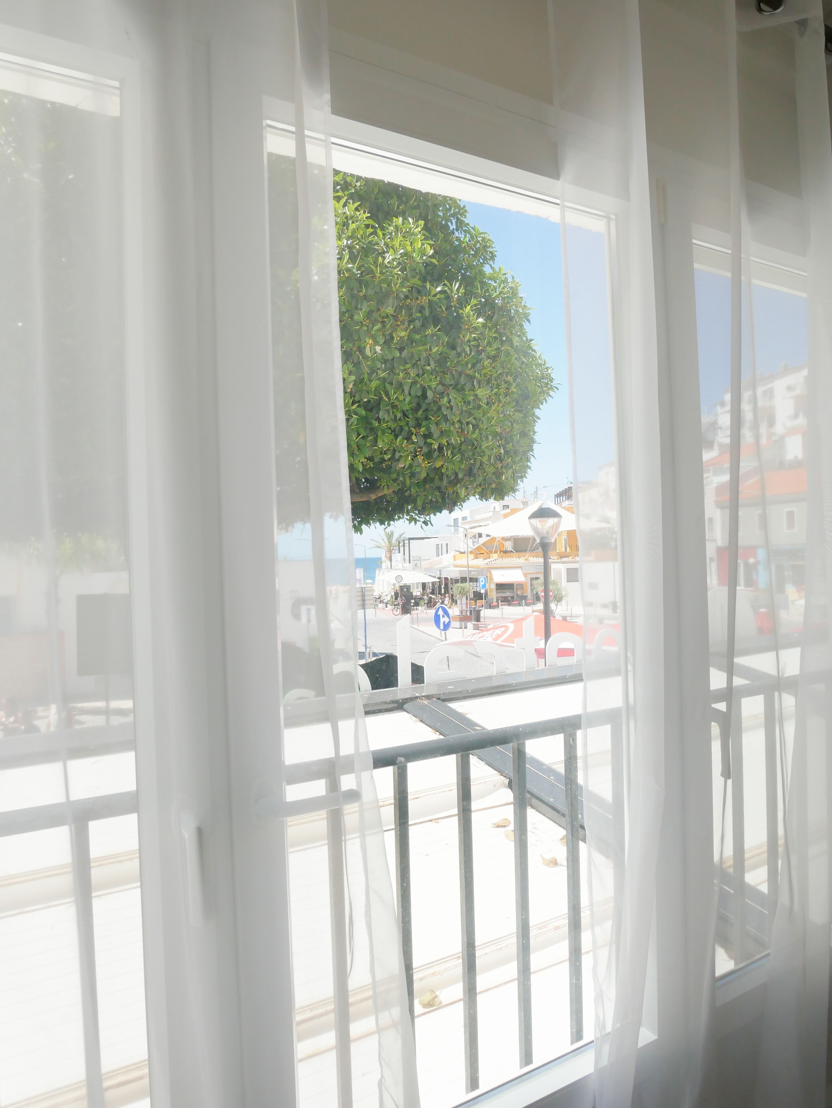 A view through a window with sheer curtains, revealing a street scene with buildings, a tree, and a pedestrian crossing.