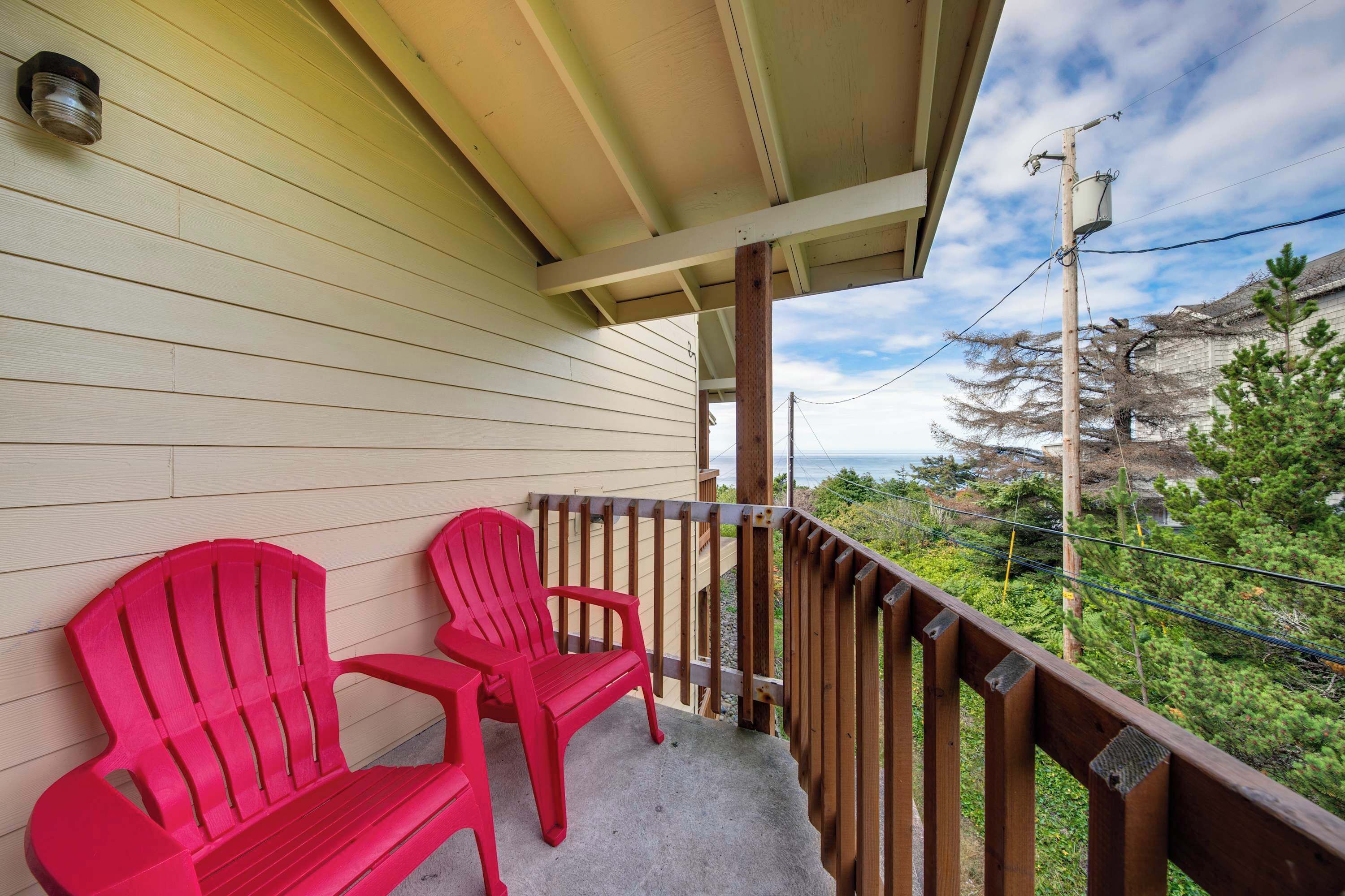 A balcony with two red Adirondack chairs, a wooden railing, and a view of the ocean and trees.