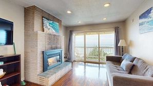 A living room with a fireplace, a sofa, a TV, and a balcony with a view. - Dog Friendly Oceanview Condo with Great Location and Nearby Beach Access (Lincoln City)