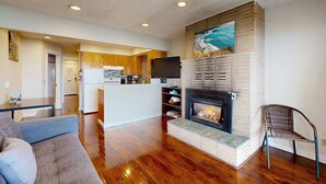 A modern living room with hardwood flooring, a fireplace, a sofa, and a kitchen in the background. - Dog Friendly Oceanview Condo with Great Location and Nearby Beach Access (Lincoln City)