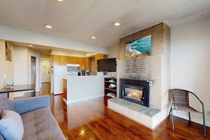 A modern living room with hardwood flooring, a fireplace, a sofa, and a kitchen in the background. - Dog Friendly Oceanview Condo with Great Location and Nearby Beach Access (Lincoln City)