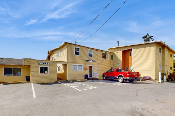 Un motel à deux étages, avec une enseigne indiquant « BUREAU » et « SAUMON », un camion rouge garé devant, et un ciel bleu dégagé.