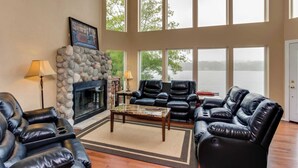 A spacious living room with a stone fireplace, leather sofas, and large windows overlooking a body of water. - Dog Friendly Lakefront Vacation Home with Stunning Views and Spacious Deck (Lincoln City)