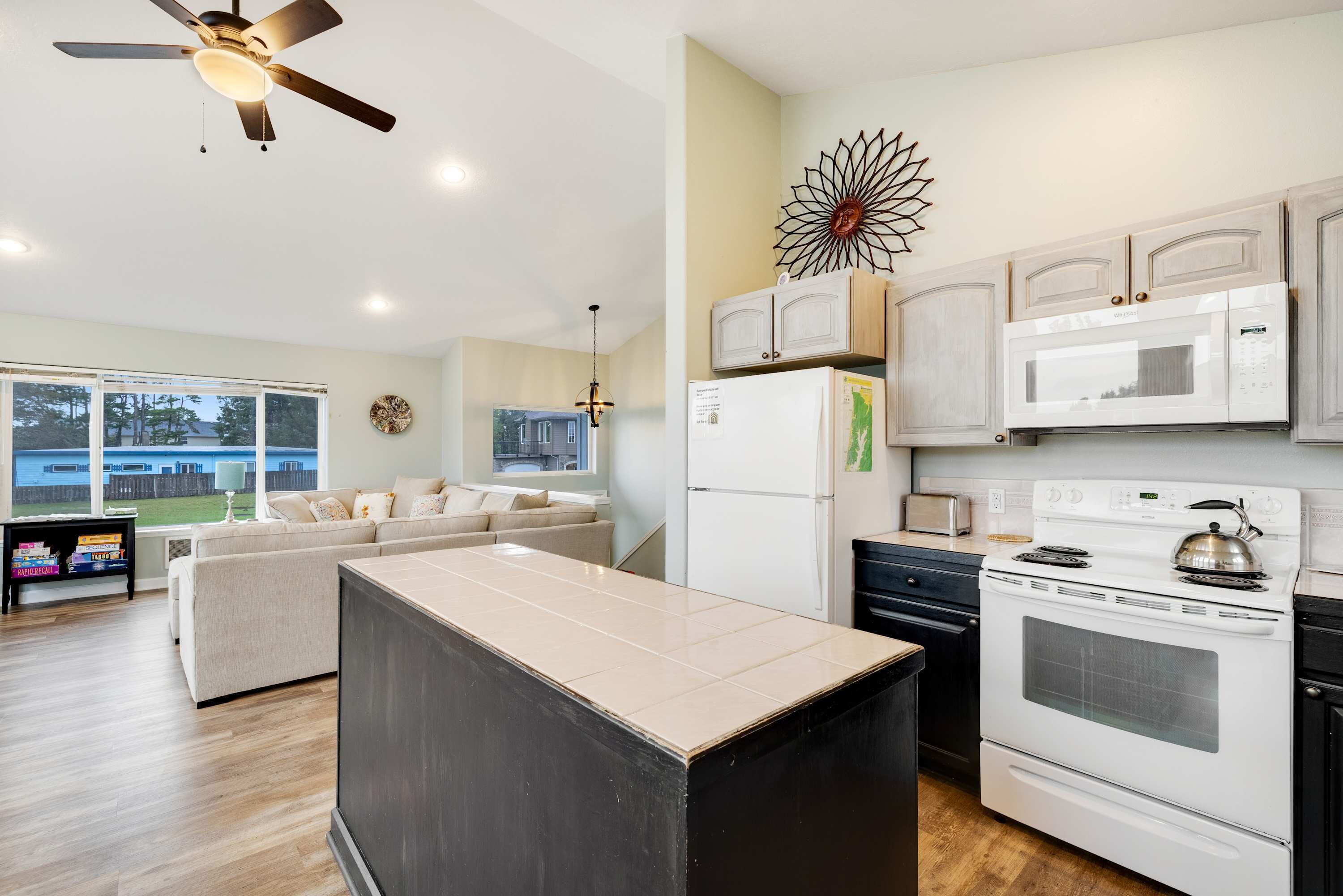 A modern kitchen with white cabinets, a built-in oven, microwave, and refrigerator. There is a large window providing natural light and a view of the outdoors.