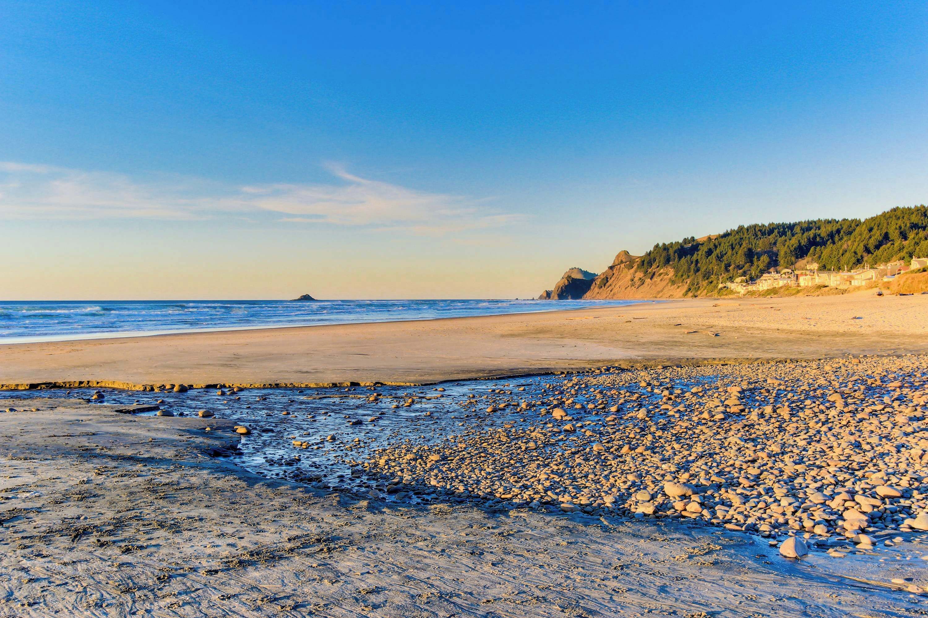 A beach with pebbles and sand, a clear sky, and a forested hill in the background.