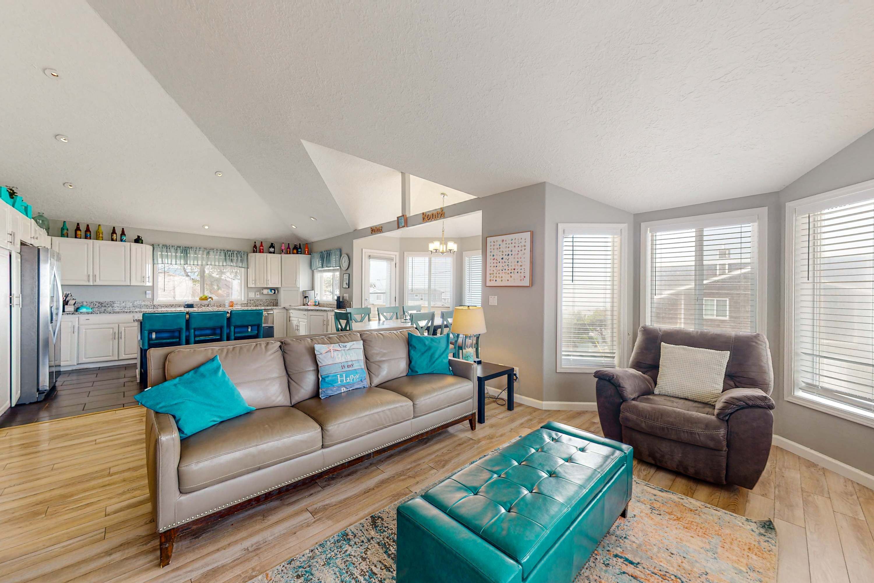 A spacious living room with a beige sofa, a teal ottoman, and a brown armchair. A kitchen is visible in the background with white cabinets and blue appliances.