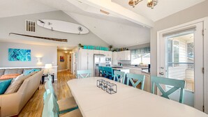 Eine moderne Küche mit weißen Schränken, ein Essbereich mit einem weißen Tisch und Türkis-Stühlen sowie ein Surfbrett an der Wand.