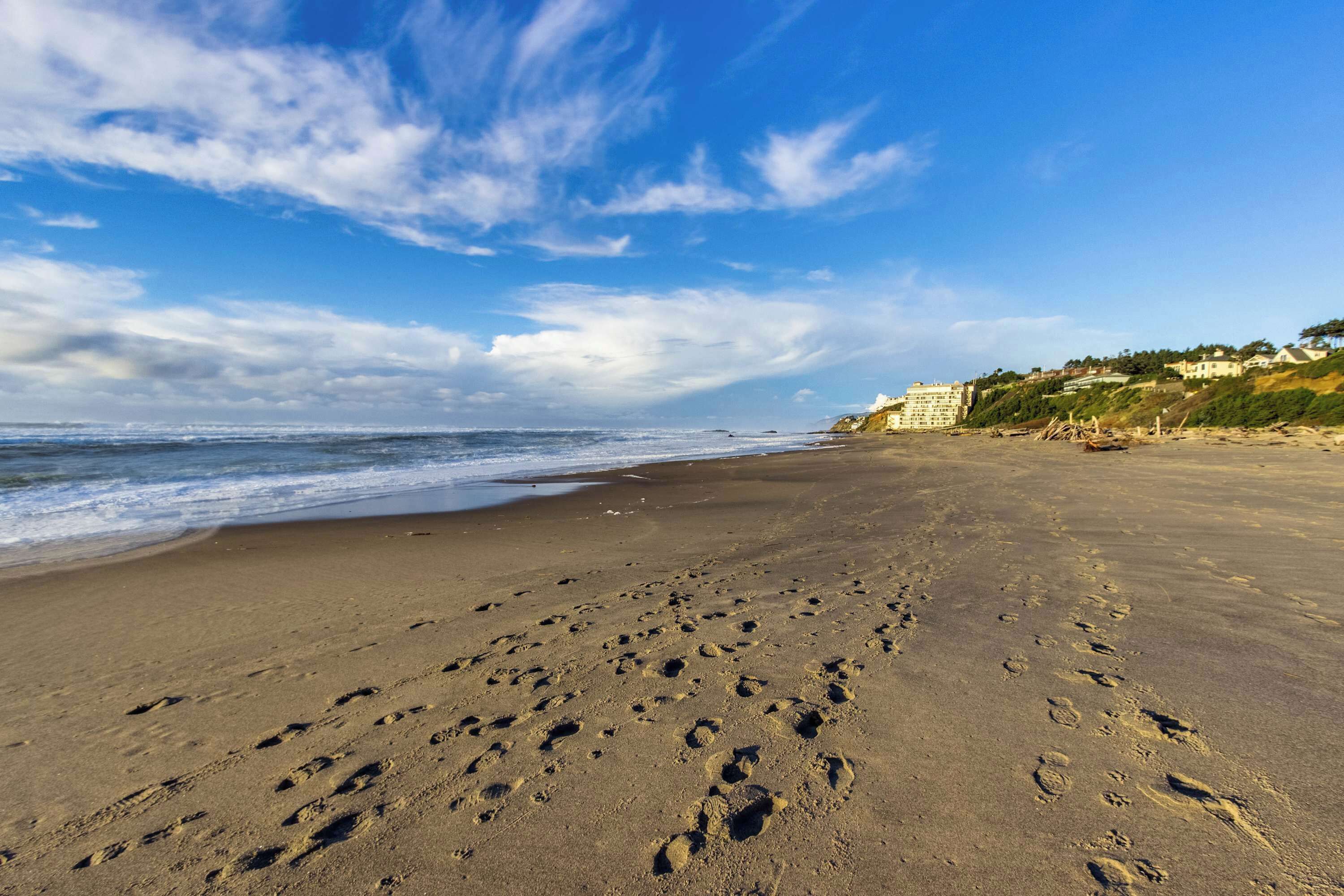 Une plage où l’on voit des empreintes de pas menant à l’eau, un ciel dégagé et des bâtiments en arrière-plan.