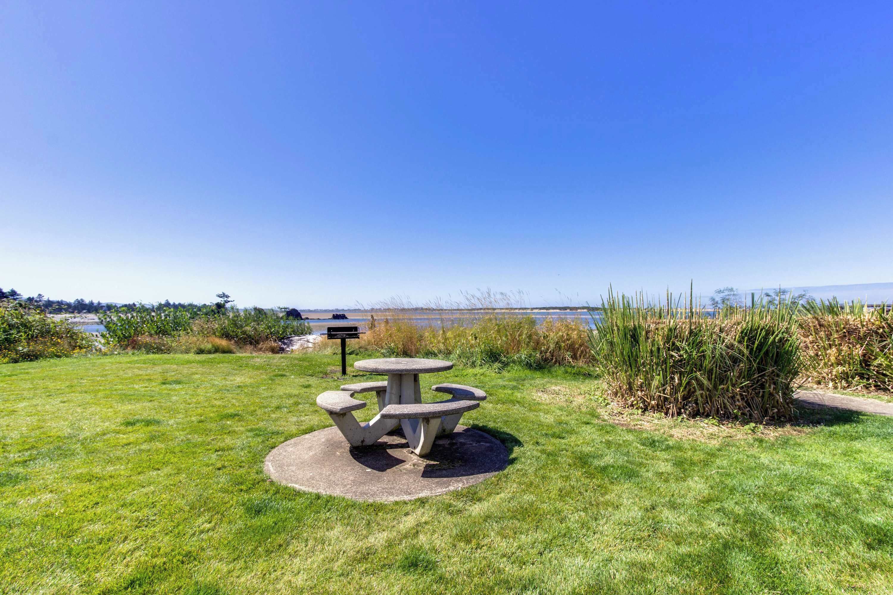 Une table de pique-nique avec un banc et un panneau, installée dans un espace verdoyant offrant une vue sur une étendue d’eau.