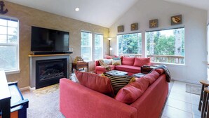 A living room with a fireplace, a large TV, a red sofa, and large windows with a view of trees. - Spacious dog friendly home with WiFi and fireplace near the beach (Lincoln City)