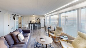 Un salon moderne avec un canapé, des fauteuils et une table basse. On aperçoit une cuisine à l’arrière-plan, avec des tabourets de bar.