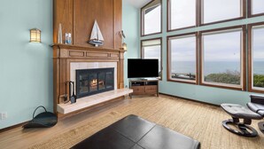 Una sala de estar con chimenea, televisor y ventanas grandes que ofrecen vista al océano.