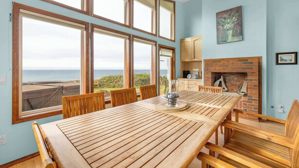 Un comedor con una amplia mesa de madera y sillas, una chimenea de ladrillo y ventanas grandes que ofrecen vistas al océano.