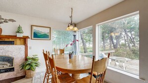 A dining area with a round wooden table, six wooden chairs, a chandelier, a fireplace, a large window with a view of trees, and a framed landscape painting on the wall. - Waterfront house with bay and ocean views large deck and quiet location (Lincoln City)