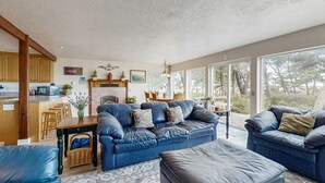A spacious living room with a blue leather sofa, a matching ottoman, and a dining area with a wooden table and chairs. Large windows provide a view of the outdoors. - Waterfront house with bay and ocean views large deck and quiet location (Lincoln City)