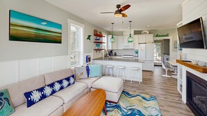A modern living room with a sofa, ottoman, and a wooden coffee table. A kitchen with white cabinets and a dining area with white stools is visible in the background. - Wonderful coastal escape in the trees with private hot tub and fast WiFi dogs OK (Lincoln City)
