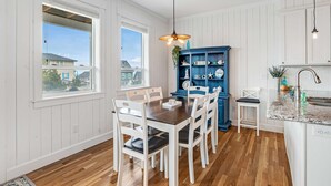 Zona de dinar amb taula de fusta i cadires blanques, un armari blau amb objectes decoratius i cuina amb armaris blancs i terra de fusta.