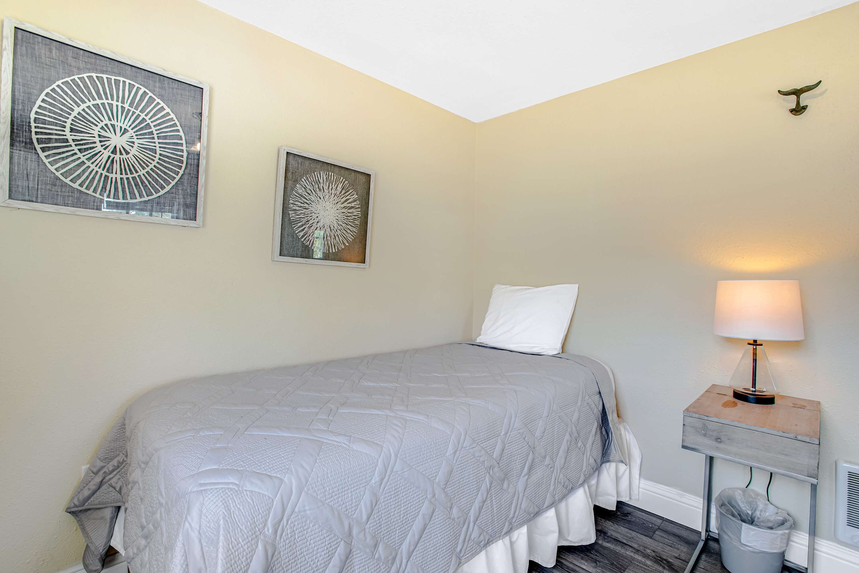 Una cama bien hecha con un edredón gris, una lámpara de noche y dos cuadros en la pared.
