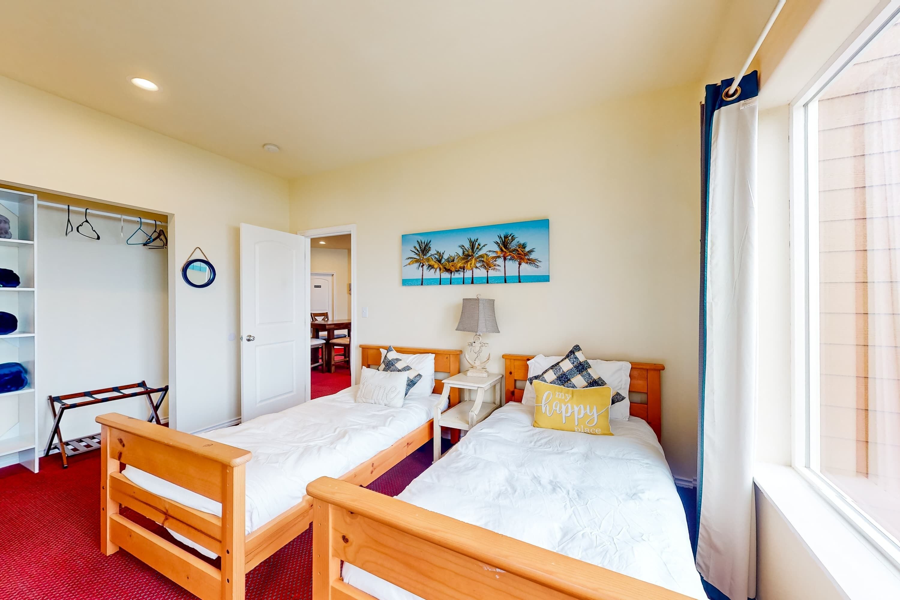 Ein Zimmer mit zwei Holzbetten, einem roten Teppich, einem Palmenmotiv an der Wand und einer weißen Tür sowie einem Fenster mit Vorhängen.