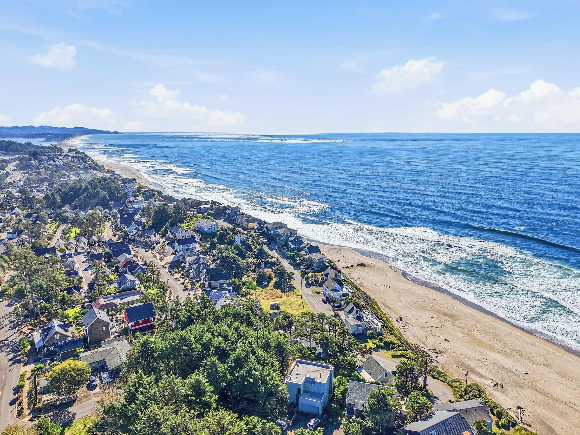 Ett flygbildsbild av ett bostadsområde vid kusten, med hus längs stranden, en klar himmel och havet i bakgrunden.