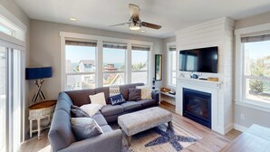 Ein modernes Wohnzimmer mit einem grauen Ecksofa, einem Kamin, einem Flachbildfernseher und großen Fenstern mit Blick ins Freie.