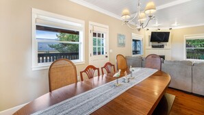 A dining area with a wooden table, chairs, and a chandelier. There are windows with views of greenery and a balcony. A television is mounted on the wall. - Family Home with Game Room WiFi Fireplace Private Hot Tub Walk to Beach (Lincoln City)
