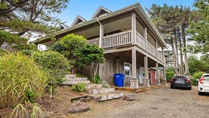 A two-story wooden house with a porch, surrounded by trees and shrubs. - Family Home with Game Room WiFi Fireplace Private Hot Tub Walk to Beach (Lincoln City)