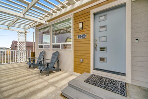 A covered porch with Adirondack chairs, a wooden door with a number plate, and a window with a warning sign. - Light filled home in the heart of town with deck and with D steps to beach access (Lincoln City)