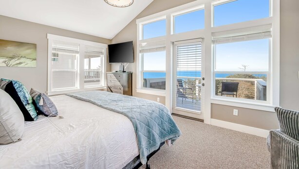 Quarto com uma cama grande, televisão e vista para o oceano através de amplas janelas.