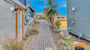 A narrow brick pathway leading to a house with a wooden door and a small garden area with a palm tree. - Ocean view home 1 block from the beach with hot tub deck and fireplace (Lincoln City)