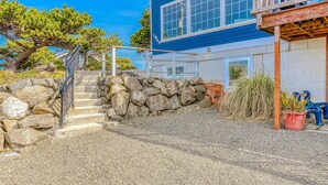 A two-story building with a stone staircase leading to the entrance, a wooden deck, and a clear blue sky. - Ocean view home 1 block from the beach with hot tub deck and fireplace (Lincoln City)