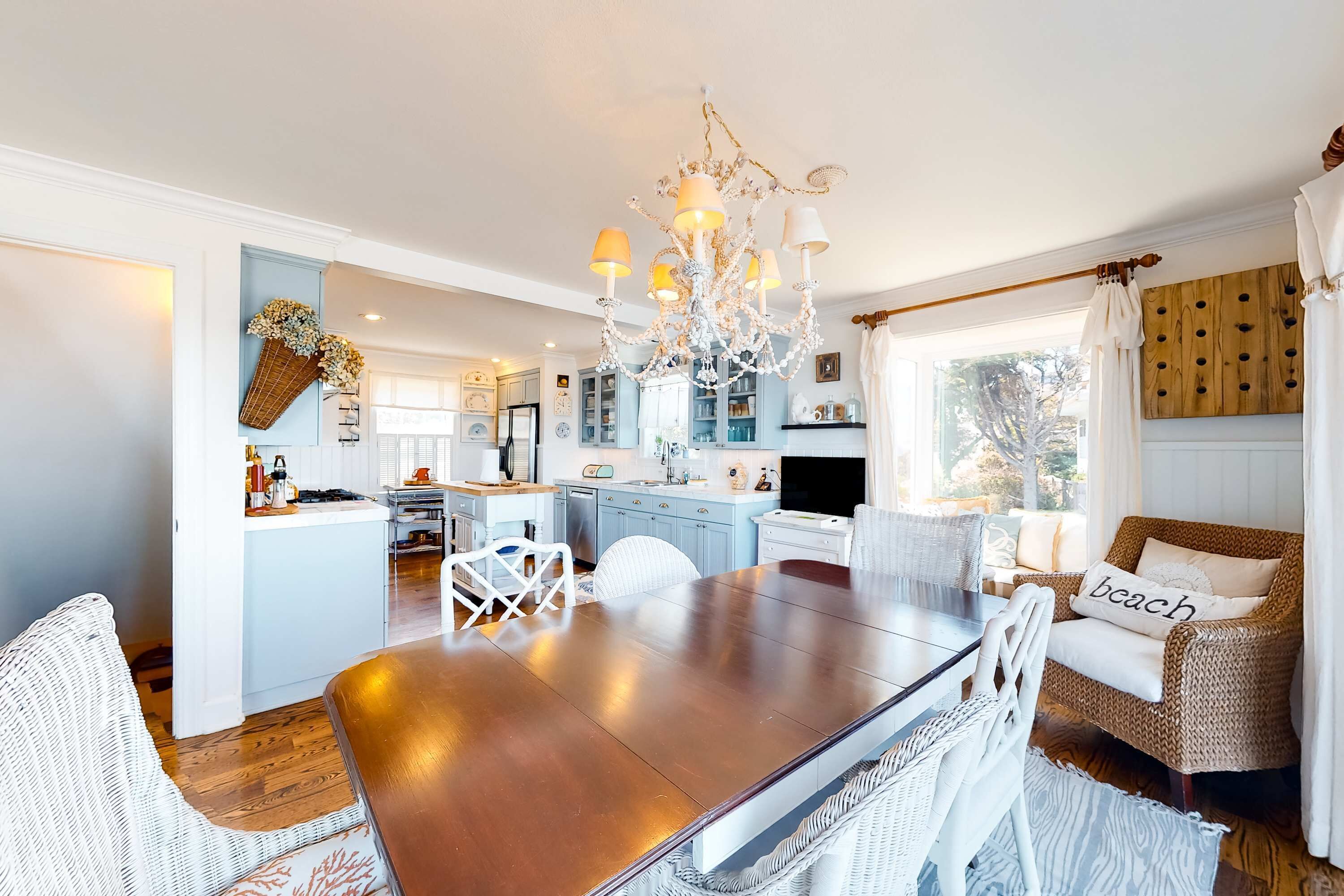 A spacious dining area with a wooden table and white chairs, a chandelier, and a kitchen in the background.