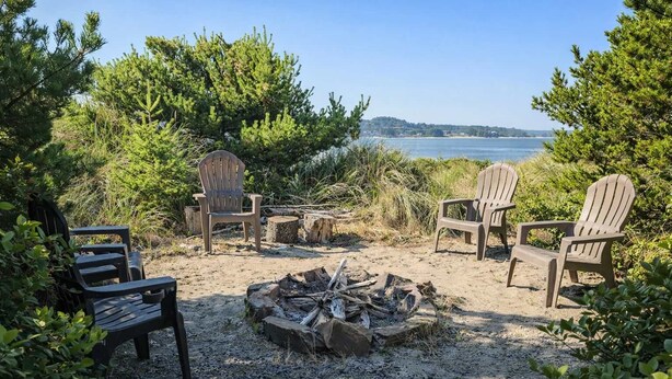 Ein Strand mit Adirondack-Stühlen und einem Feuerkorb, umgeben von Grün und Blick aufs Meer.
