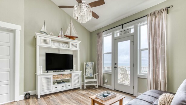 A living room with a television, white entertainment unit, wooden floor, grey sofa, and a balcony door. - Ocean view home with hot tub game room furnished deck and fireplaces (Lincoln City)
