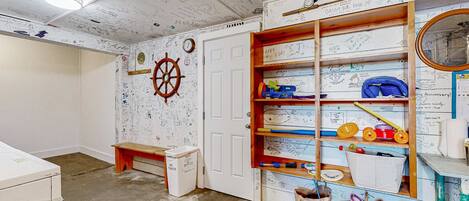 A room with a white door, a wooden shelf with various items, a wall clock, and a ship's wheel decoration.