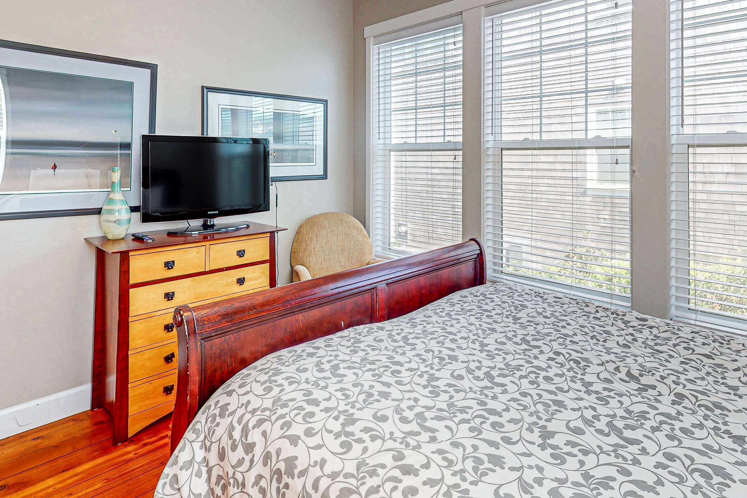 Une chambre à coucher avec un lit, un téléviseur posé sur une commode en bois et de grandes fenêtres équipées de stores.