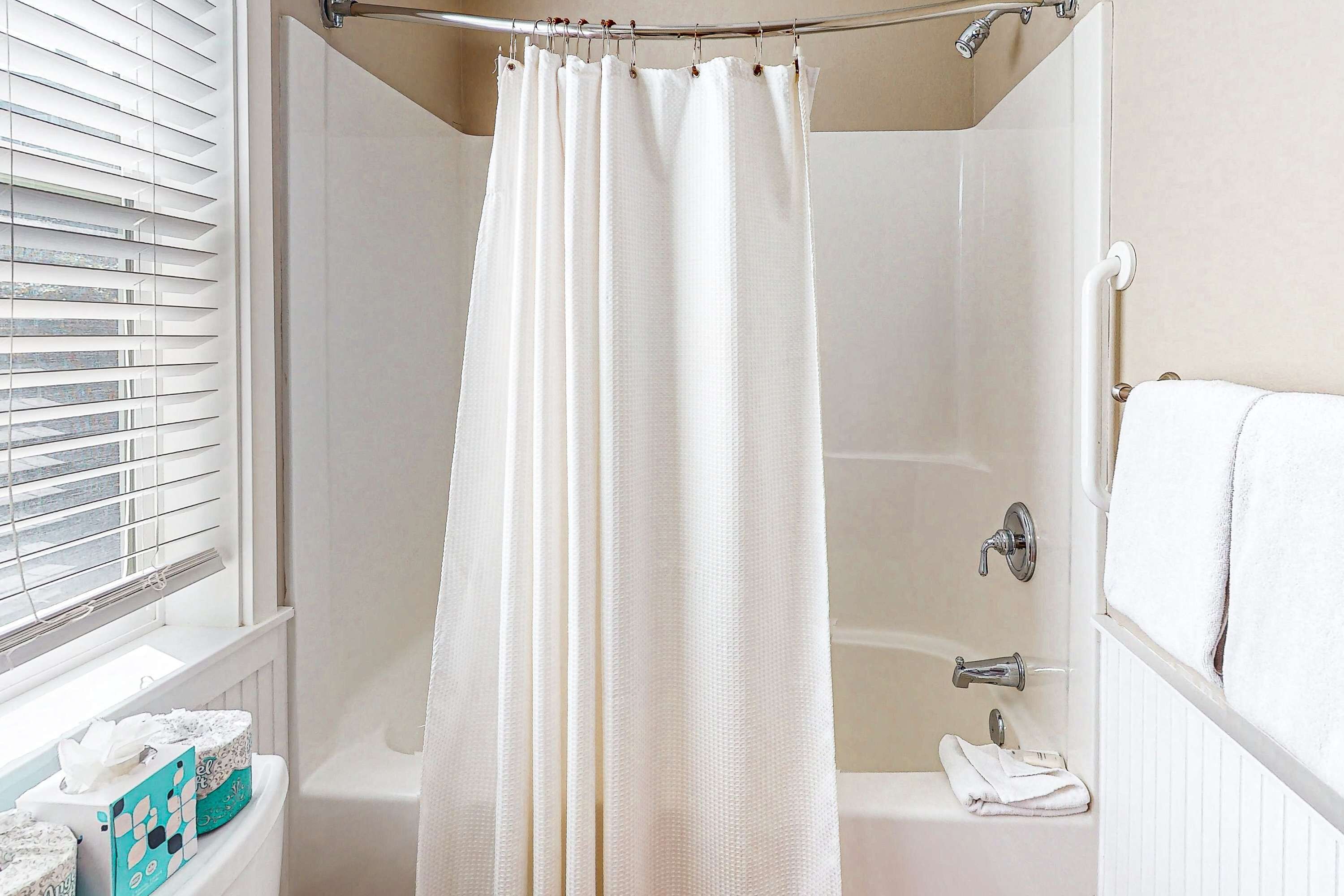 A bathroom with a shower curtain, a bathtub, a towel rack, and a window with blinds.