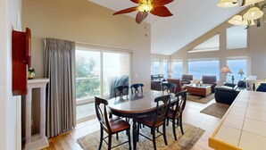 Un vaste espace de vie comprenant une table à manger ronde, plusieurs chaises, un ventilateur de plafond et de grandes fenêtres offrant une vue sur l’extérieur.